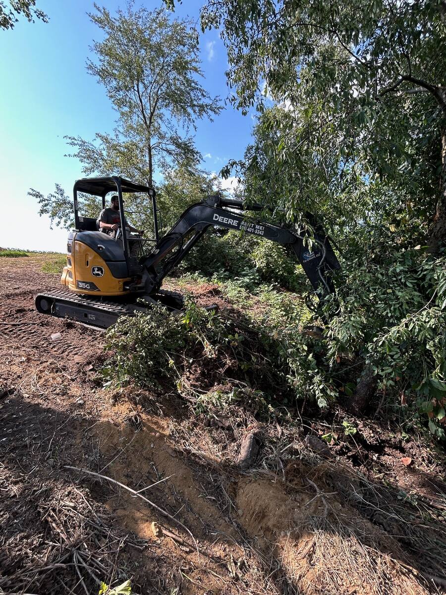 Mini excavator clearing brush along a tree line to prep ground for a food plot